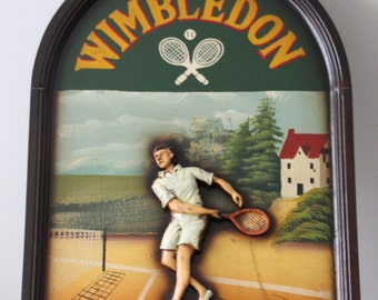 May include: Wooden framed wall art featuring a tennis scene. The top arch reads "WIMBLEDON" in yellow letters. A tennis player is depicted in white attire, mid-swing. The background shows a tennis court, trees, and a house.