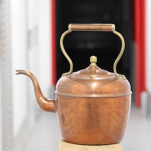 Puede incluir: Una tetera de cobre con asa y boquilla de latón. La tetera tiene un cuerpo redondeado y una tapa con un pequeño pomo de latón. El asa se curva hacia arriba y luego hacia abajo hacia los lados de la tetera.