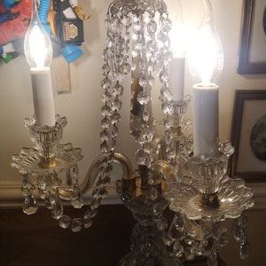 May include: A crystal candelabra with three lit candle-shaped bulbs. The lamp features a decorative base, curved arms, and hanging crystal accents. The light fixture is a vintage style, with a classic design.