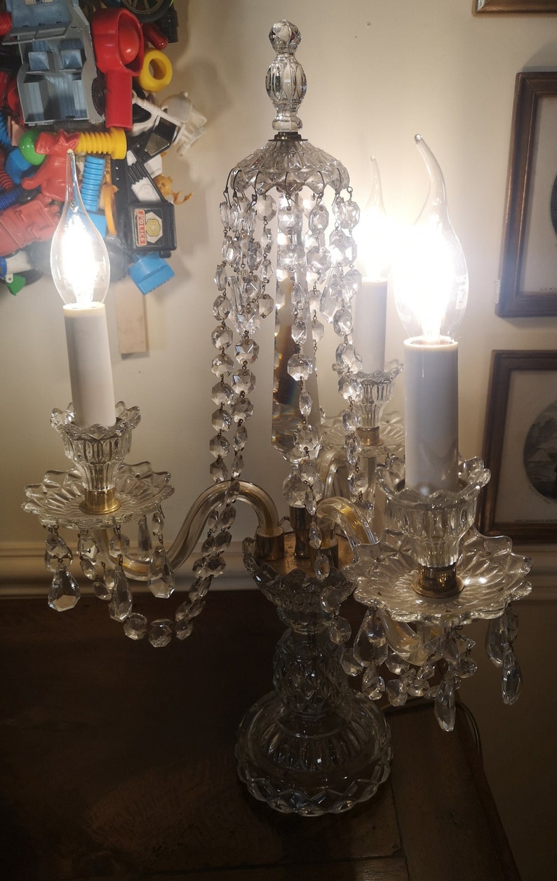 May include: A crystal candelabra with three lit candle-shaped bulbs. The lamp features a decorative base, curved arms, and hanging crystal accents. The light fixture is a vintage style, with a classic design.