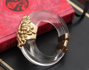 May include: A clear plastic bangle bracelet with a gold-toned, ornate, floral design on one side. The bracelet is open and has a hinged closure.