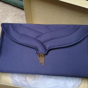 May include: A navy blue clutch with a quilted flap and a gold-colored chain tassel. The rectangular bag is in an open cardboard box with tissue paper. The clutch has a satin finish.