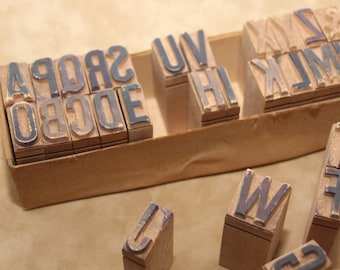 Puede incluir: Una caja de madera vintage llena de sellos de letras, incluyendo las letras de la A a la Z. Los sellos son de madera y tienen una almohadilla de tinta azul oscuro. La caja es de cartón y es de color marrón.