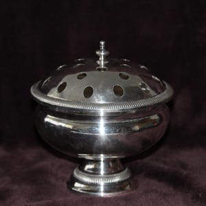 Può includere: Un portafiori argentato con coperchio rotondo perforato e base a piedistallo. Il coperchio ha fori multipli per disporre i fiori. Il contenitore ha un bordo decorativo e una piccola punta in cima. Probabilmente un oggetto vintage.