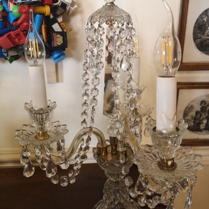 May include: A clear glass chandelier with crystal detailing and two light bulbs. The chandelier features a decorative base and arms, with hanging crystal beads. The light bulbs are candle-shaped. The chandelier is on a dark brown surface.