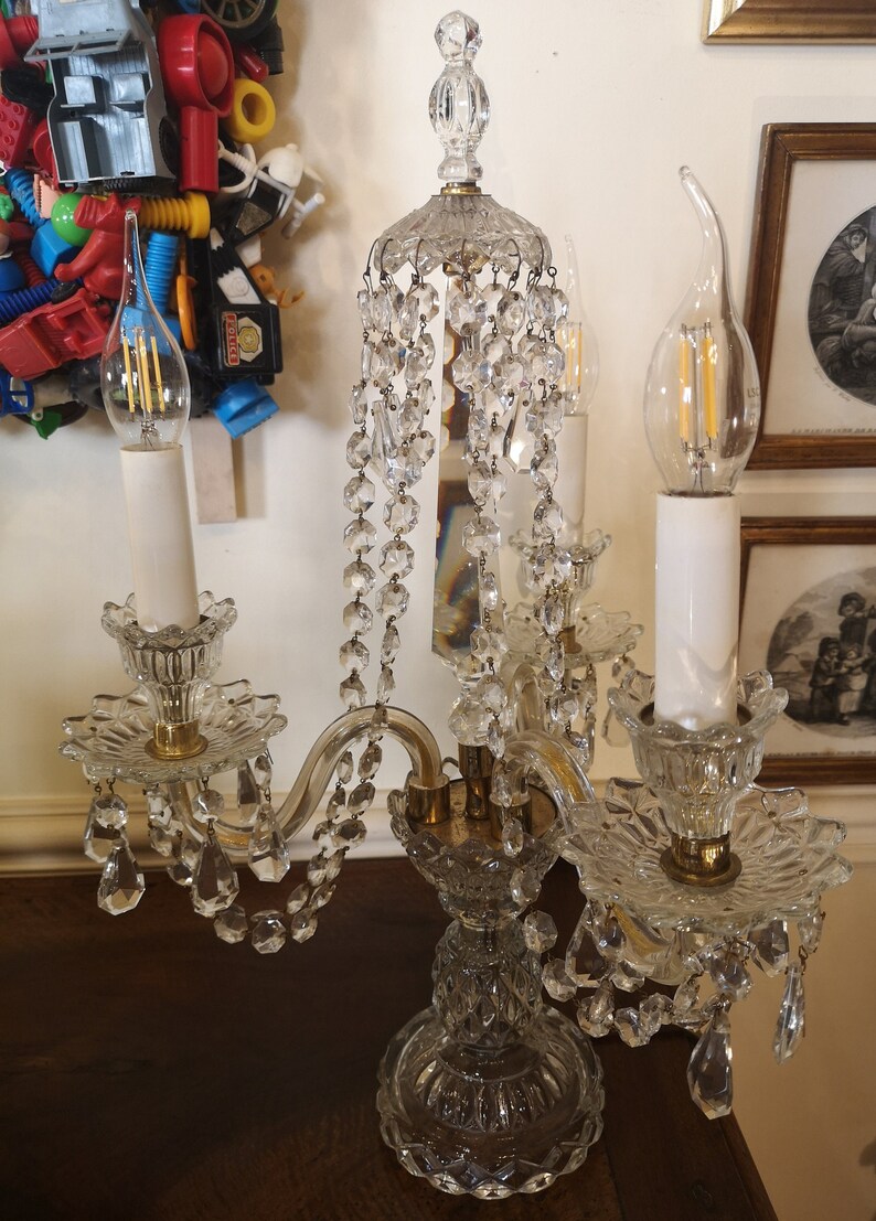 May include: A clear glass chandelier with crystal detailing and two light bulbs. The chandelier features a decorative base and arms, with hanging crystal beads. The light bulbs are candle-shaped. The chandelier is on a dark brown surface.