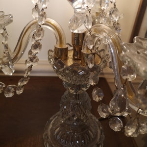May include: A clear glass candelabra with crystal detailing. The candelabra features a decorative base and curved arms. Crystal droplets are suspended from the arms. The candelabra is on a dark wooden surface.