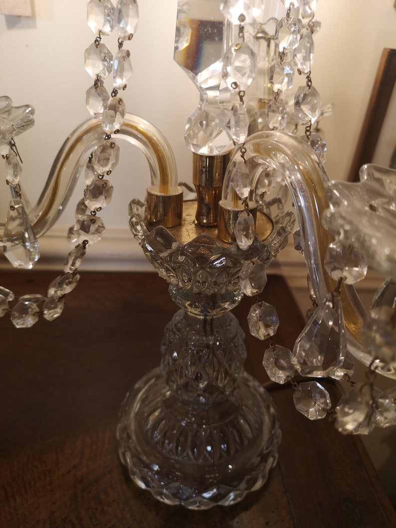 May include: A clear glass candelabra with crystal detailing. The candelabra features a decorative base and curved arms. Crystal droplets are suspended from the arms. The candelabra is on a dark wooden surface.