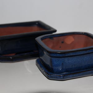 May include: Two blue ceramic bonsai pots with matching trays. The pots are square with a dark blue glaze and a reddish-brown interior. The trays are the same colour as the pots. The pots are sitting on a white surface.