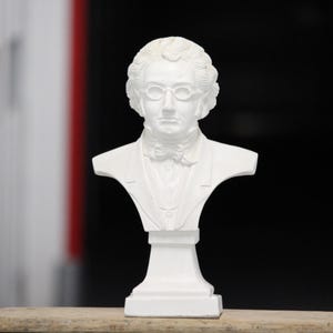 May include: A white bust sculpture of a man with curly hair, glasses, and a bow tie. The sculpture is on a pedestal base and is set against a blurred background.