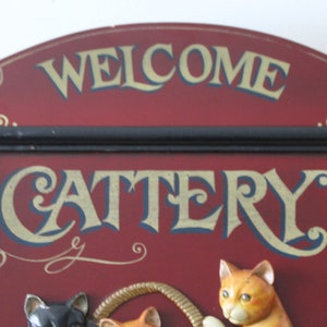 Może przedstawiać: Drewniany znak w kolorze czerwonym z czarną ramą i złotymi napisami, na kt&oacute;rym widnieje napis "Welcome Cattery". Znak przedstawia tr&oacute;jwymiarowe koty bawiące się sznurkiem.