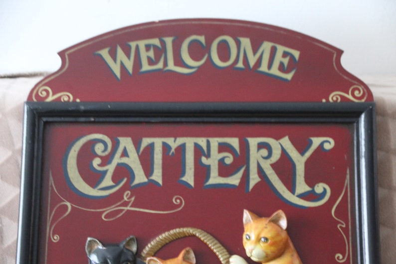 Może przedstawiać: Drewniany znak w kolorze czerwonym z czarną ramą i złotymi napisami, na kt&oacute;rym widnieje napis "Welcome Cattery". Znak przedstawia tr&oacute;jwymiarowe koty bawiące się sznurkiem.