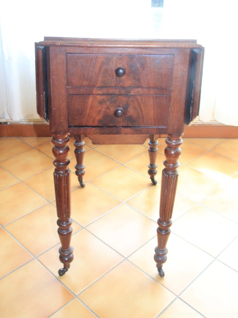 Puede incluir: Una mesa auxiliar de madera oscura con dos cajones y patas torneadas. La mesa tiene una esquina redondeada y se apoya en cuatro ruedas.