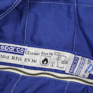 May include: Close-up of a blue Sparco racing suit. The suit features white stitching, a black zipper, and a label with the Sparco logo, model information, and size 58. The suit also has safety certifications.
