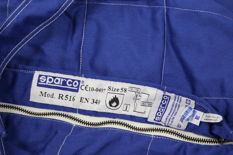 May include: Close-up of a blue Sparco racing suit. The suit features white stitching, a black zipper, and a label with the Sparco logo, model information, and size 58. The suit also has safety certifications.