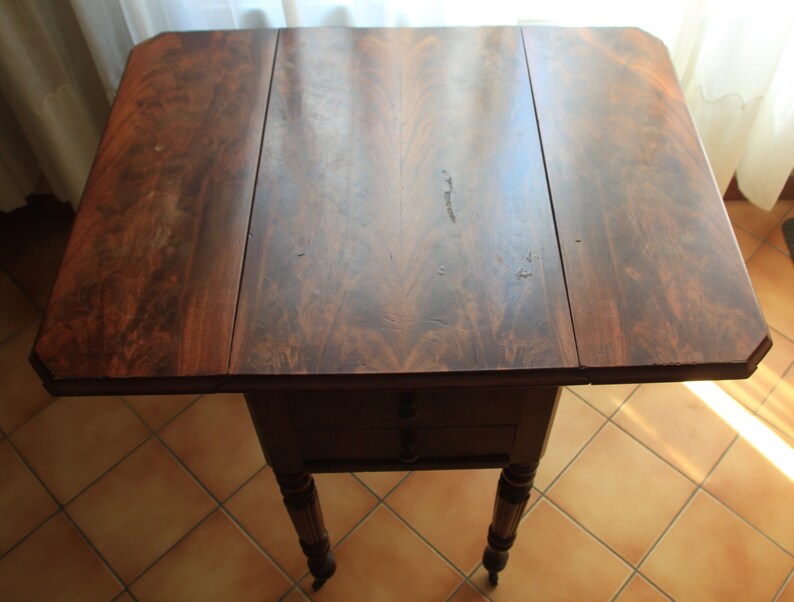 Puede incluir: Una mesa de madera marr&oacute;n oscuro con un dise&ntilde;o de hoja ca&iacute;da. La mesa tiene dos cajones y cuatro patas torneadas. La mesa est&aacute; en buen estado y tiene una hermosa p&aacute;tina.