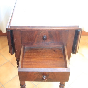 Puede incluir: Una mesa de madera oscura con tapa abatible, dos cajones y patas torneadas. La mesa tiene una tapa blanca y est&aacute; sobre un suelo de baldosas.