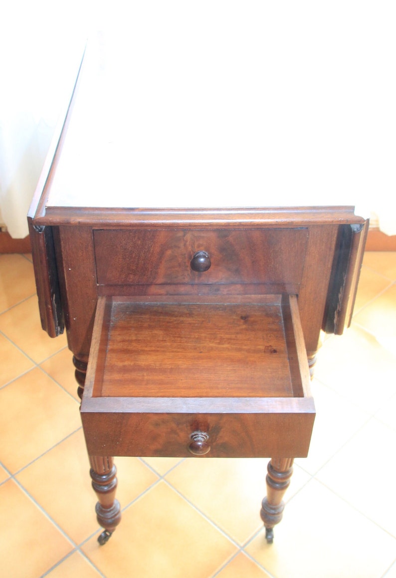 Puede incluir: Una mesa de madera oscura con tapa abatible, dos cajones y patas torneadas. La mesa tiene una tapa blanca y est&aacute; sobre un suelo de baldosas.