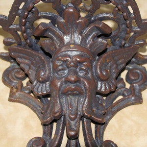 May include: Close-up of a cast iron wall decor piece featuring a detailed face with wings and a beard. The brown metal has an aged, rustic appearance, with intricate scrollwork and a decorative top.