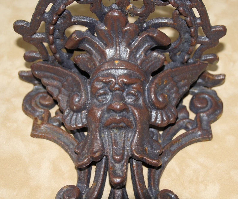 May include: Close-up of a cast iron wall decor piece featuring a detailed face with wings and a beard. The brown metal has an aged, rustic appearance, with intricate scrollwork and a decorative top.