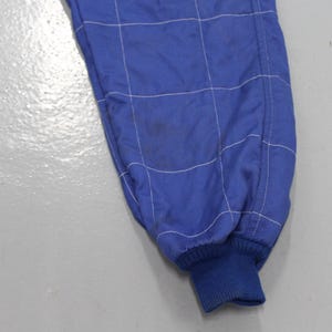 May include: Close-up of a blue racing suit leg with white stitching in a grid pattern. The cuff is a darker blue ribbed knit. There is a stain on the fabric. The suit appears to be made of a lightweight material.
