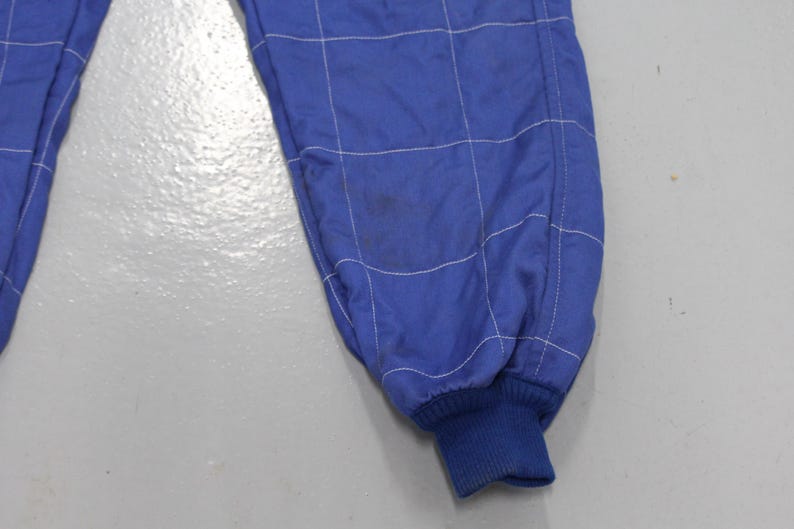 May include: Close-up of a blue racing suit leg with white stitching in a grid pattern. The cuff is a darker blue ribbed knit. There is a stain on the fabric. The suit appears to be made of a lightweight material.