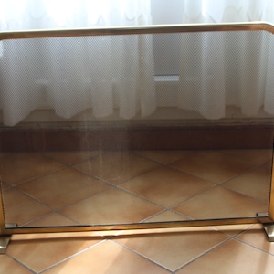 May include: A rectangular fireplace screen with a gold-colored metal frame and a fine mesh screen. The screen has a rounded top and stands on two rectangular feet. The screen is designed to protect against sparks and embers.