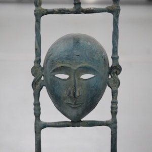 May include: A blue-green metal sculpture featuring a central human face mask with almond-shaped eyes and a neutral expression. The mask is framed by a vertical metal structure with twisted side bars and horizontal crossbars, showing a weathered finish.