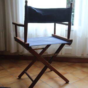 May include: A wooden director's chair with a blue canvas seat and back. The chair is folded and standing on a tiled floor.