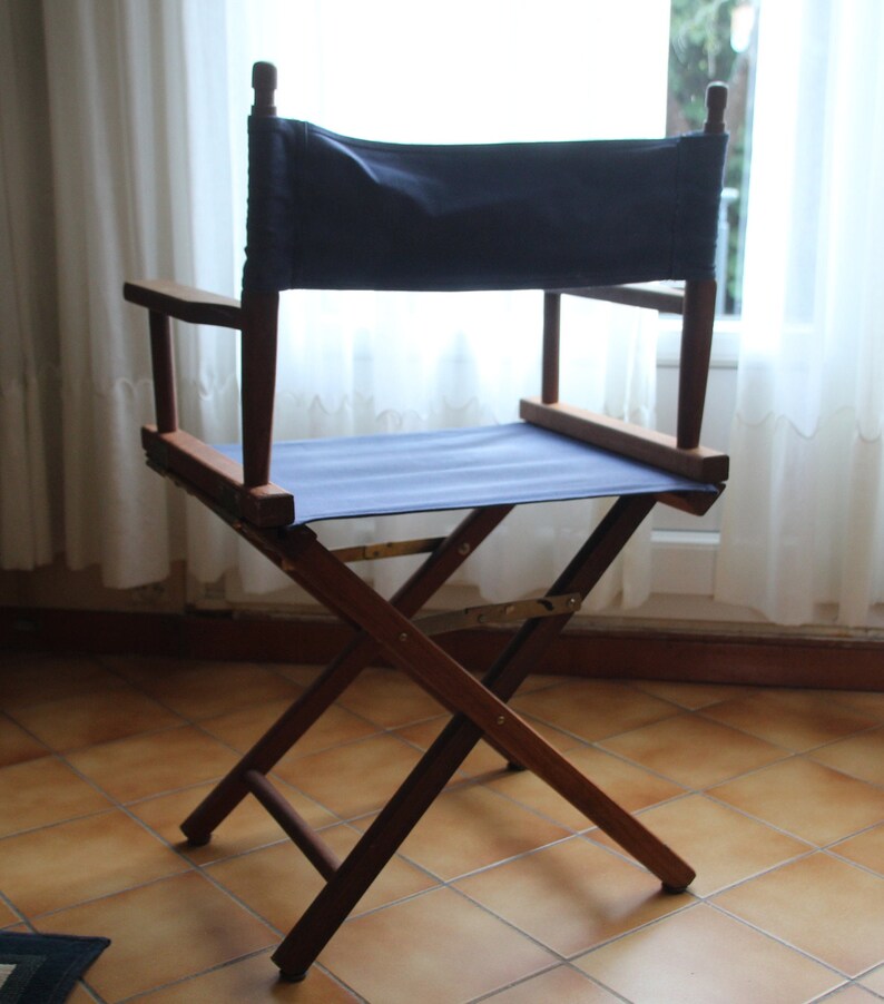 May include: A wooden director's chair with a blue canvas seat and back. The chair is folded and standing on a tiled floor.