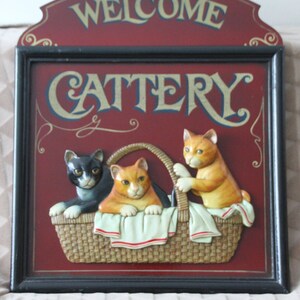 Może przedstawiać: Drewniany znak z napisem "Welcome Cattery" złotymi literami na czerwonym tle. Trzy kocięta są przedstawione w wiklinowym koszu, jedno kocię czarno-białe i dwa pomarańczowe kocięta.