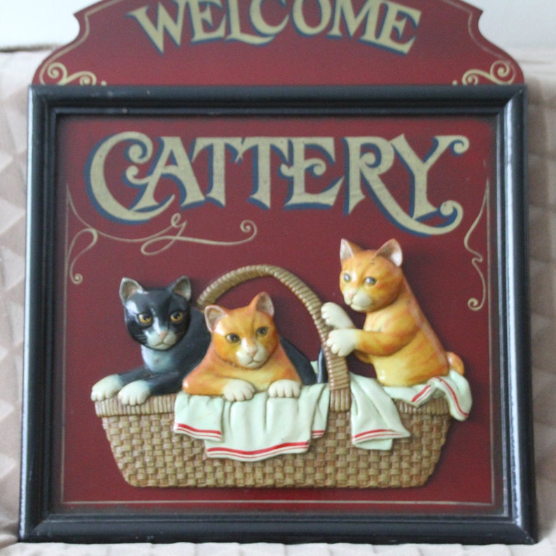 Może przedstawiać: Drewniany znak z napisem "Welcome Cattery" złotymi literami na czerwonym tle. Trzy kocięta są przedstawione w wiklinowym koszu, jedno kocię czarno-białe i dwa pomarańczowe kocięta.