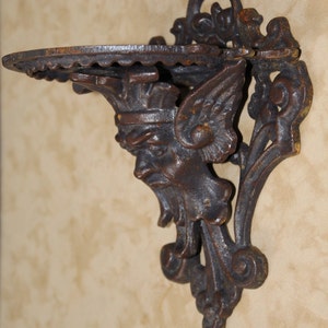 May include: Ornate, dark brown cast iron wall shelf. The shelf features a detailed face with wings and a crown. The shelf has a scalloped edge and is mounted on a decorative bracket. The shelf is designed to hold small items.