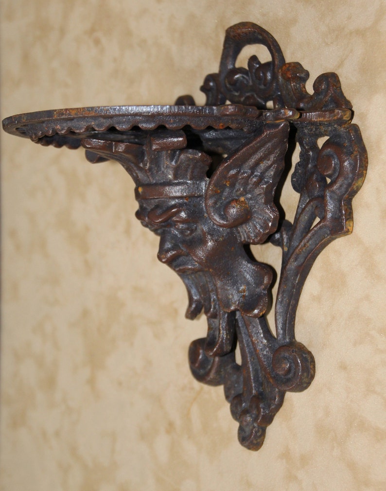 May include: Ornate, dark brown cast iron wall shelf. The shelf features a detailed face with wings and a crown. The shelf has a scalloped edge and is mounted on a decorative bracket. The shelf is designed to hold small items.