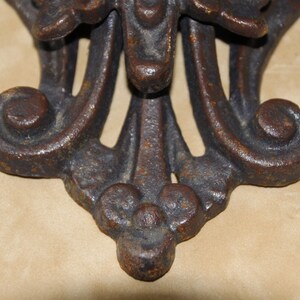 May include: Close-up of a decorative, ornate metal object with a dark, aged finish. The design features symmetrical, stylized floral or leaf motifs, with a central element and curved, scroll-like details. The object appears to be made of cast iron.
