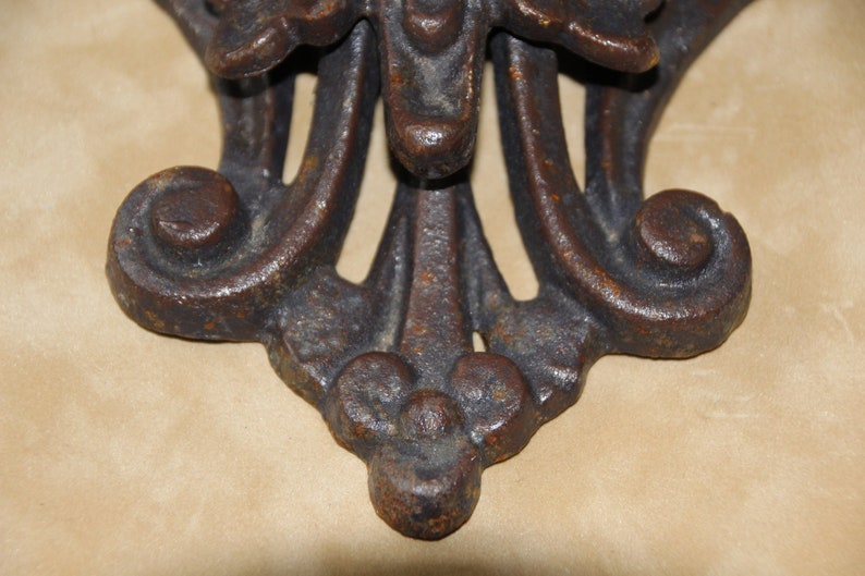 May include: Close-up of a decorative, ornate metal object with a dark, aged finish. The design features symmetrical, stylized floral or leaf motifs, with a central element and curved, scroll-like details. The object appears to be made of cast iron.