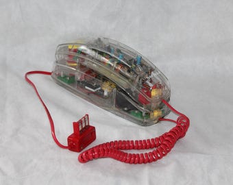 Może przedstawiać: Przezroczysty telefon z widocznymi elementami wewnętrznymi. Telefon ma czerwony, zwinięty przewód słuchawkowy i czerwoną podstawę. Konstrukcja prezentuje wewnętrzne obwody i części elektroniczne.