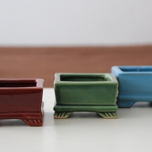 May include: Three small, square ceramic bonsai pots. The pots are in shades of red-brown, green, and blue. Each pot has a rectangular shape with a flared rim and decorative feet. The pots are empty and sit on a white surface.