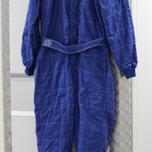 May include: A blue racing suit with white stitching and a matching belt. The suit has long sleeves and legs, with the brand name "Sparco" visible on the shoulders. The suit is hanging on a wooden hanger.