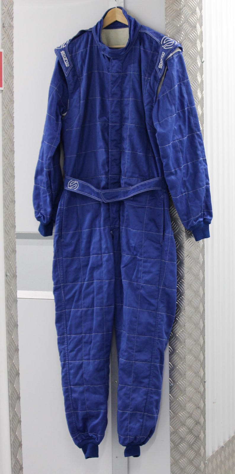 May include: A blue racing suit with white stitching and a matching belt. The suit has long sleeves and legs, with the brand name "Sparco" visible on the shoulders. The suit is hanging on a wooden hanger.