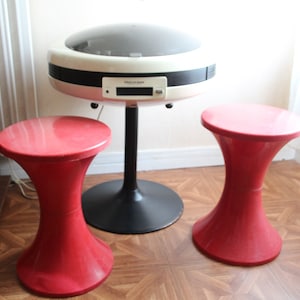 Op de afbeelding: Een witte en zwarte retro-platenspeler met een rond, koepelvormig deksel staat op een zwarte standaard met een brede basis. Twee rode plastic krukjes met witte bases staan voor de platenspeler.