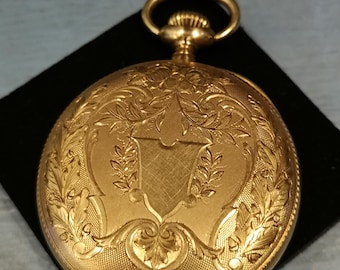 May include: A gold pocket watch with an ornate engraved design on the front. The watch has a hinged lid and a loop for a chain.
