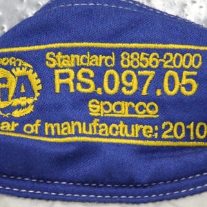 May include: Close-up of a blue fabric with yellow embroidered text and a logo. The text includes "Standard 8856-2000", "RS.097.05", "Sparco", and "Year of manufacture: 2010". The logo is a yellow FIA emblem.