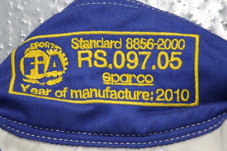 May include: Close-up of a blue fabric with yellow embroidered text and a logo. The text includes "Standard 8856-2000", "RS.097.05", "Sparco", and "Year of manufacture: 2010". The logo is a yellow FIA emblem.