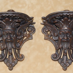 May include: Pair of antique-style, cast iron wall sconces. Each sconce features a detailed, weathered finish and a grotesque face design with wings and ornate scrollwork. The sconces are designed to hold small items.