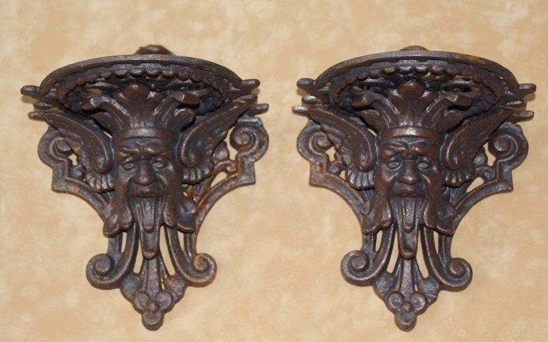 May include: Pair of antique-style, cast iron wall sconces. Each sconce features a detailed, weathered finish and a grotesque face design with wings and ornate scrollwork. The sconces are designed to hold small items.