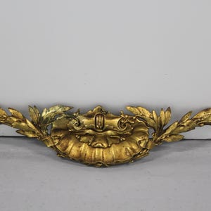 May include: Ornate gold-colored decorative furniture hardware. The piece features a central shell-like design with leaf accents extending outwards. The hardware is likely made of metal and has a detailed, aged appearance.