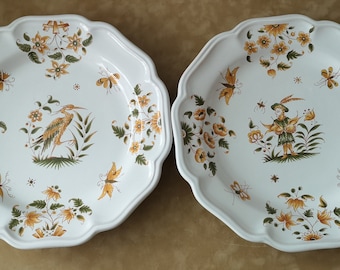 Pode incluir: Dois pratos de cerâmica branca com bordas festoneadas. Os pratos apresentam um design floral com acentos amarelos e verdes. Um prato tem um design de pássaro, enquanto o outro tem uma figura de uma pessoa segurando uma flor.
