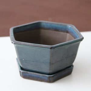 Op de afbeelding: Een zeshoekige keramische plantenbak in een gedempte teal kleur met een bijpassende schotel. De plantenbak heeft een glanzende afwerking en een donker interieur. De randen hebben een donkerdere blauwe tint. Geschikt voor kleine planten.