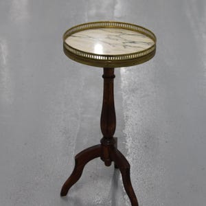 Peut inclure: Une petite table d'appoint ronde avec un plateau en marbre et un bord en laiton, sur trois pieds en bois. La table est sur un sol gris.